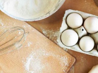 Bowl of flour, eggs, whisk, and an apron on a wooden surface